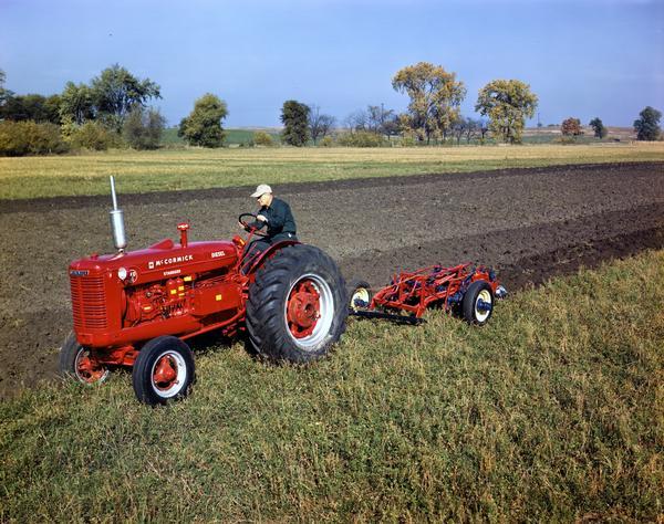 Three-quarter view towards front left of a farmer plowing a field with a McCormick WD-6 tractor and an attached plow.