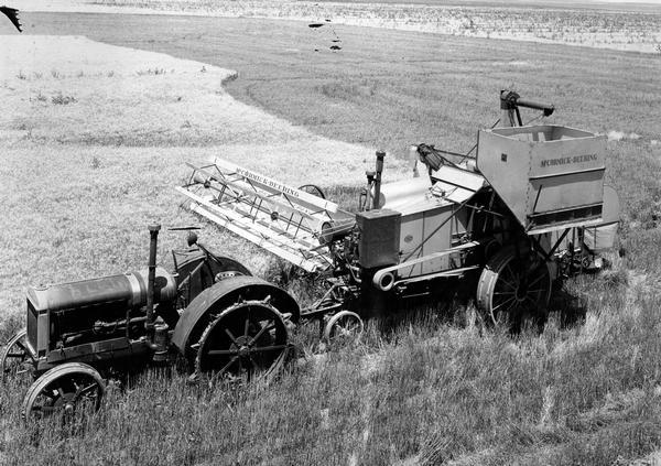 Elevated view of a McCormick-Deering No. 41 harvester-thresher (combine) hitched to a tractor in a field. Original caption reads: "While the no. 41 is not yet in full production, it is expected to replace the no. 11 harvester-thresher. It is a 3-wheel in-line machine equipped with 16-foot folding-type platform, five-section straw rack, recleaner, and platform power control. The latter device permits the tractor operator to raise or lower the platform as desired by merely pulling one of two ropes conveniently attached to the tractor."