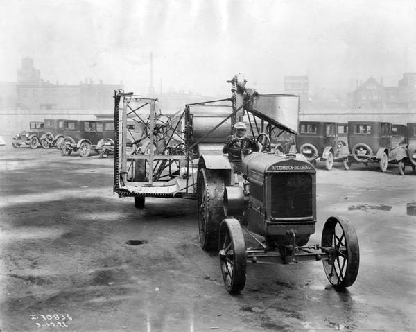 Worker transporting a McCormick-Deering No. 8 power-drive harvester-thresher (combine) with a McCormick-Deering tractor. The harvester-thresher is equipped with a domestic type of grain tank and with platform folded for transport. Automobiles are parked in the background.