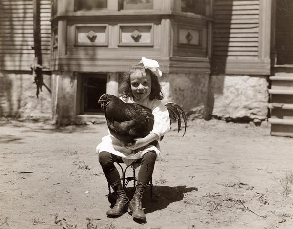 Portrait of a young girl sitting on a stool holding a rooster in her lap, in front of Sunnyside School. The girl was participating in a third year "living things" program at the school.