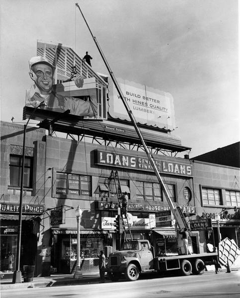 Construction workers erecting a billboard for Hines Lumber using a crane mounted on the back of an International heavy-duty truck. The billboard is sitting atop a line of shops at the corner of Milwaukee and Ashland streets.