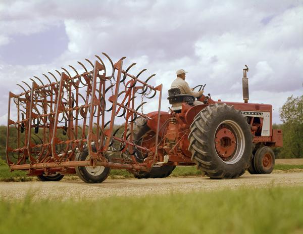 View towards a farmer operating a Farmall 806 tractor with attached spring harrow along a gravel road.