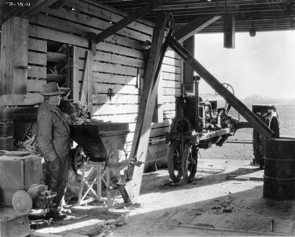 Farmer grinding corn in a barn with a belt-driven McCormick-Deering feed grinder powered by a Farmall tractor.