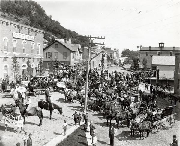 Elevated view of people, horses, and wagons gathered in the center of Fountain City to celebrate McCormick Day. Scherer's Hotel is at left. The event was organized by a McCormick Harvesting Machine Company dealer to publicize the delivery of new machines to customers.