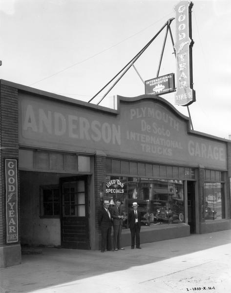Men standing in front of Anderson Garage, an International truck dealership. The men are (left to right): W.E. Penrod, salesman; Neils Anderson, manager; and A.W. Sorenson, salesman. Signs on the dealership building advertise: "Plymouth, DeSoto, International trucks", "Goodyear" and "Used Car Specials".
