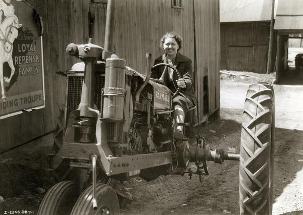 Mrs. Lucile Stronsky, manager of Savanna Farm Supply Company, sitting on a McCormick-Deering Farmall F-12 tractor outside the Mt. Connell  Farm Supply store. A portion of an advertising poster for the "Loyal Repenski Family" riding troupe is on the wall of the building.