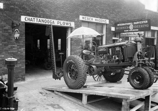 Farmall F-20 tractor with attached mower and sun shade on a ramp outside a McCormick-Deering farm equipment dealership. Original caption reads: "The Farmers Truck and Tractor Company, Selma, Alabama, use their loading ramp to display a Farmall tractor in front of their building. Such a display throws the tractor about four feet from the ground - high enough to be seen over other equipment displayed and a sure way to draw attention of the passerby." Signs on the dealership building also advertise "Chattanooga Plows" and "Weber Wagons."