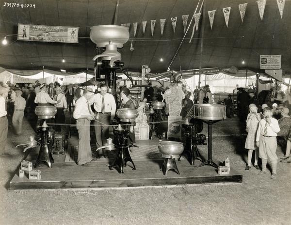 Men, women, and children at an International Harvester cream separator exhibit at the Iowa State Fair. A company representative appears to be pointing out the features of the separator to a man and woman as children are standing nearby eating ice cream cones. The display includes advertising posters, a giant cream separator, and cardboard cut-outs of a man and child working with a cream separator.