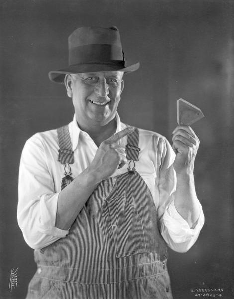 Advertising photograph of a man in overalls and hat pointing to an  International Harvester part. The photograph was taken in a studio and used in company advertising for International Harvester parts.