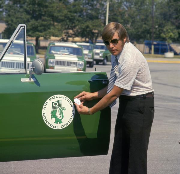 Color photograph of an air pollution inspector wearing sunglasses is holding his badge near the door emblem on his International Scout II pickup. The truck was one of a fleet of trucks used by the Air Pollution Control District of Jefferson County.