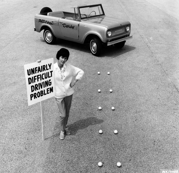 Press release photograph of a woman holding a picket sign reading "unfairly difficult driving problem" in mock protest of the difficulty of events involved in the "Petticoat Derby." Original caption reads: "This pouting picket has discovered the 'straight line problem' to be negotiated by 'Petticoat Derby' contestants isn't half as easy as it may look. The problem is one of four competitive driving maneuvers the wives of state truck driving champs will tackle in Kansas City, Missouri, September 19 at the National Truck Rodeo sponsored by the American Trucking Associations, Inc. The official derby vehicle, an International Scout 800, must have its right wheels driven between two lines of teed up balls only four inches further apart than the width of a tire."