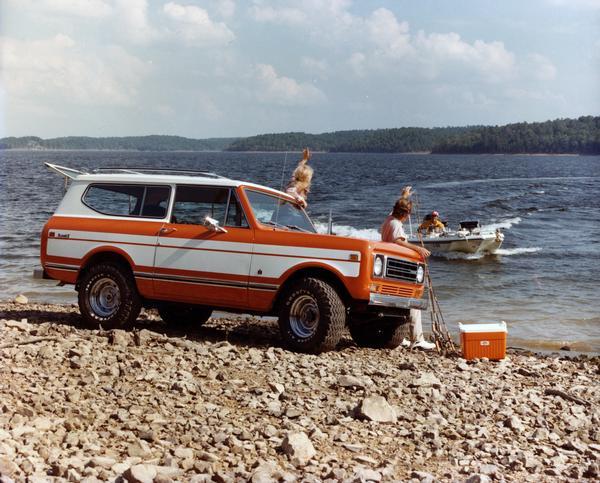 Color advertising photograph of a man and woman with their fishing gear in front of their Scout II truck on the shore waving to a man in a boat.