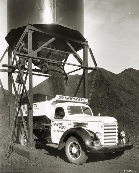 Man loading coal from an overhead bin into an International K-6 dump truck owned by South Chicago Coal and Dock Company. The slogan "coal stoker heat is best" is written over the cab of the truck.