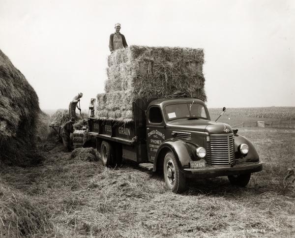 Three farm workers loading straw from a baler onto the back of an International K-6 truck owned by Sowash Grain Company.