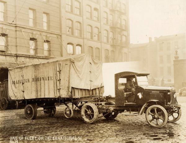 Man sitting in an International model 94 truck owned by Shapleigh Hardware. The truck is hitched to a canvas covered trailer, and is parked on a wet cobblestone street in an urban area.