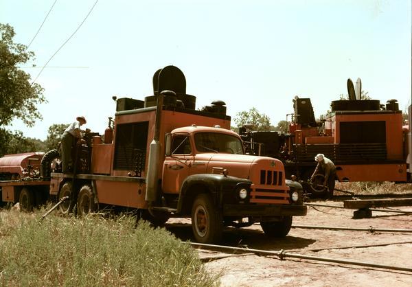Color photograph of two men servicing oil field equipment. An International model RDF-192 truck owned by "Dowell" is parked in the foreground.