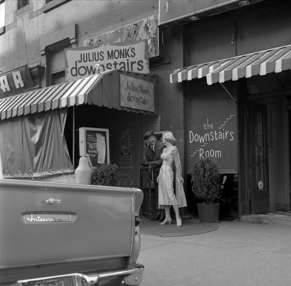 View towards a woman in fancy evening dress speaking with a doorman or chauffeur outside Julius Monk's "Downstairs Room" nightclub. The tailgate of an International truck is in the foreground.