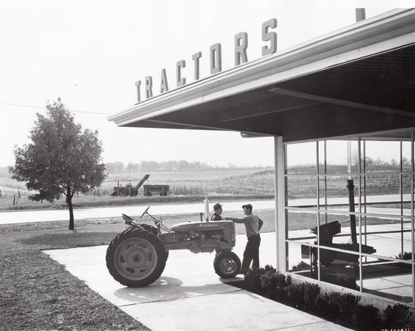 Sales agent showing a farmer a new McCormick Farmall C tractor along the storefront of an International Harvester dealership. A McCormick Farmall tractor equipped with a corn-picker and wagon is harvesting corn in the background.