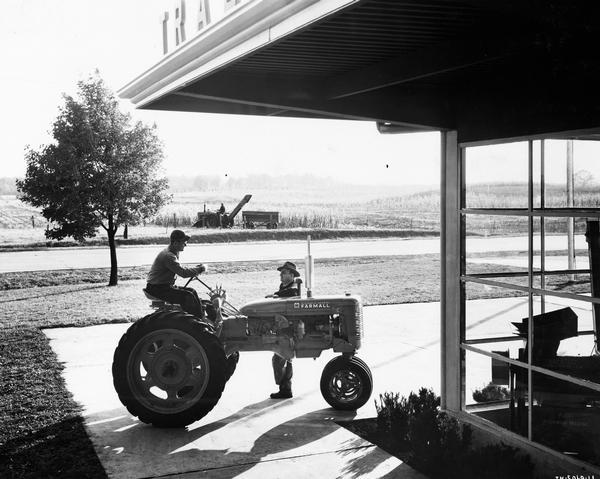 Man sitting on a Farmall C tractor in front of a prototype International Harvester dealership. The man is talking with another man. Farm fields are in the background.