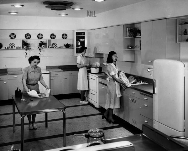 Three women in the home economics model kitchen at International Harvester's Evansville Works. Zelma Purchase is wrapping a food product at the table. Loris Knoll is cooking at a stove. And Mrs. Ethel Jean Mitchell is washing dishes at a sink. The kitchen also includes an International Harvester refrigerator and a Model 4 FC International Freezer.