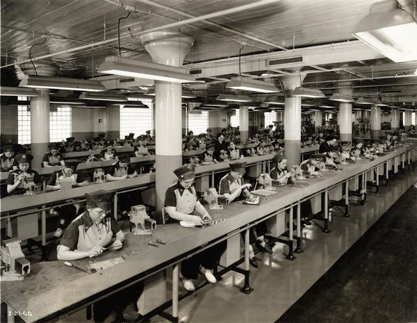 Female workers assembling torpedoes for the U.S. Military at an International Harvester factory. Original caption reads: "This large group of women employes is engaged in assembling hundreds of small, delicate pieces that go into a single part of the torpedo. They work at specially constructed benches under brilliant lights, some of them handling parts so small that they must use magnifying glasses in their work."