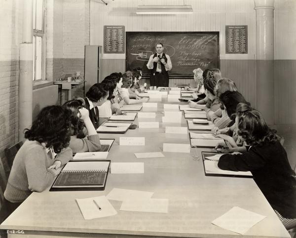 A group of new female factory workers receiving their first day of training at an International Harvester factory. The factory was  responsible for making aerial torpedoes for the U.S. Military. The women were becoming acquainted with basic mathematical terms used in torpedo production and gaining an understanding of shop tools.