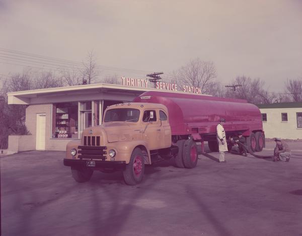 Color photograph of an International Roadliner diesel truck delivering oil to the Thrifty Service station. The delivery crew is hooking up a hose to the trailmobile oil tanker.