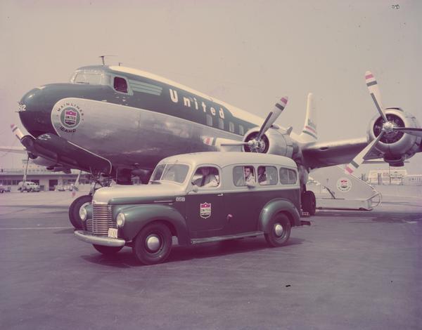 View towards men in an International KB-2 truck, with a special coach type panel body owned by United Air Lines, parked in front of a United passenger airplane.