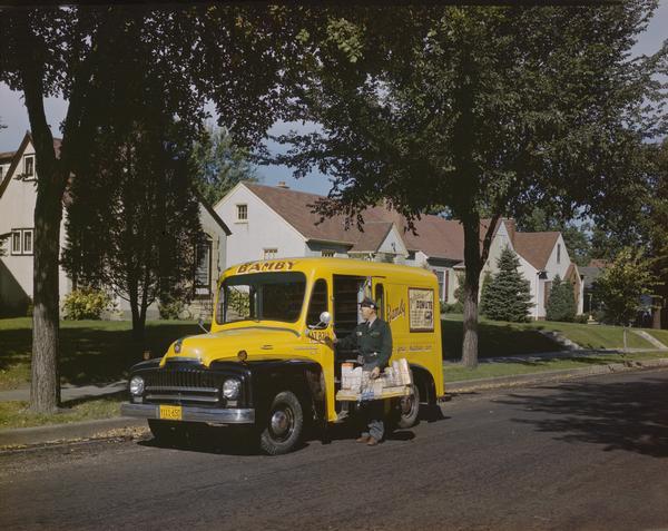 Color photograph of a man delivering Bamby brand baked goods to a residential neighborhood. The truck is one of twenty-five International LB-110 trucks operated by Excelsior Baking Co. of Minneapolis. This LB-110 was equipped with sixteen steel racks for bread and a space 12-inches high beneath the racks for other goods.