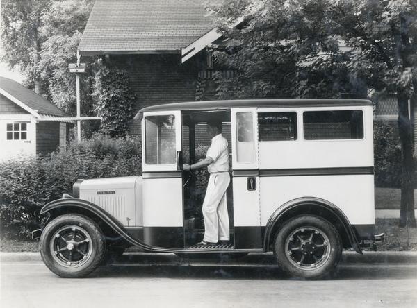 Deliveryman standing while operating an International M-2 truck on a residential street. The driver's side door is open.