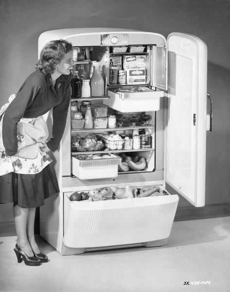 Woman wearing an apron and high heels gazing upon a well-stocked International Harvester 8H3 Deluxe refrigerator.