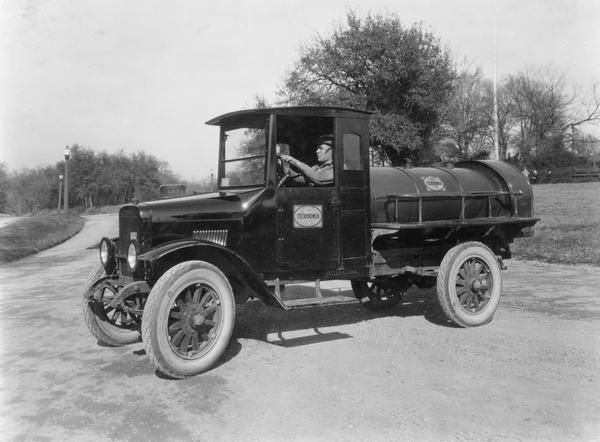 Man sitting behind the wheel of an International Model S truck with an oil tank mounted on the back. The truck belonged to the Texhoma Oil and Refinery Company of Waco, Texas.