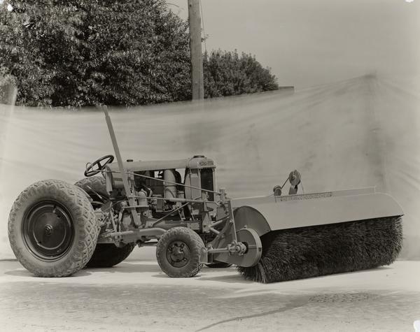 McCormick-Deering industrial tractor with an attached road sweeper. The sweeper was manufactured by the Frank G. Hough Company. A backdrop is hanging in the background.