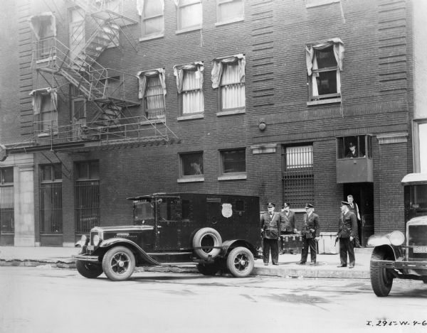 International armored truck built for Brinks Express Company parked in front of a building with armed guards. Two of the guards are holding firearms. A man wearing a suit vest and necktie is standing at the open door of the building looking out, and a man wearing a dark uniform and hat is sitting in an observation cage mounted above the door to keep watch.