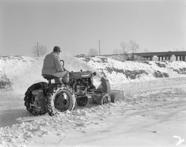 A man driving a Farmall Cub equipped with a Danco snowblower clears a road of snow.