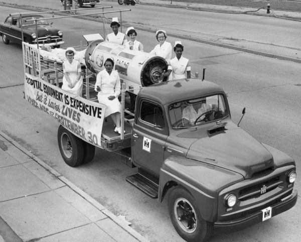 Six nurses ride in the back of an International K-160 truck, along with an iron lung and a dummy in traction. The banner on the side of the truck reads: "Hospital equipment is expensive, but it saves lives.  Vote Yes September 30."