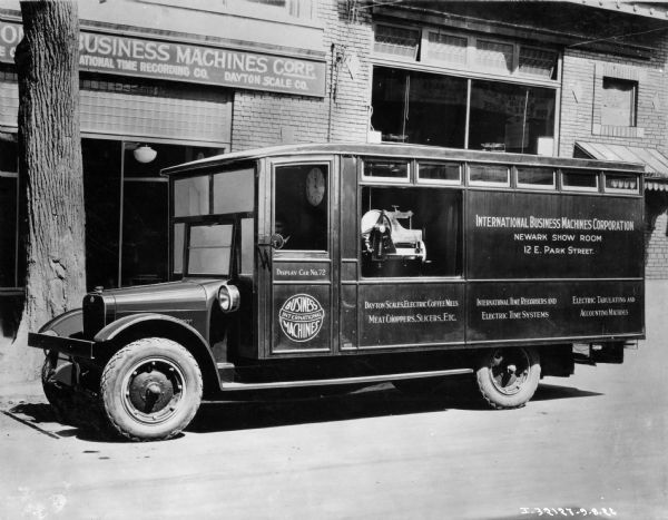 International Business Machines (IBM) display truck built by International Harvester. The truck is parked on a city street and bears the text: "Dayton Scales, Electric Coffee Mills, Meat Choppers, Slicers, Etc., International Time Recorders and Electric Time Systems, Electric Tabulating and Accounting Machines." There is a clock on the wall inside the cab.