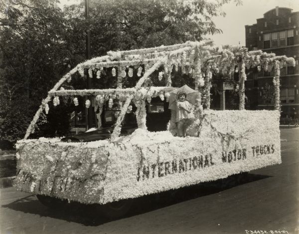 Little girl in a frilly dress posing with an umbrella while standing on a flower-covered parade float. The float is built around an International truck and bears the words: "International Motor Truck."