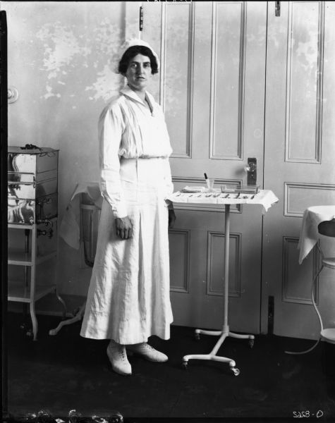 Nurse standing beside a tray of medical instruments in a medical office at one of International Harvester's factories. The nurse is dressed in a white uniform, including a cap and lace-up boots. The factory is most likely Osborne Works (later known as Auburn Works) in Auburn, New York.