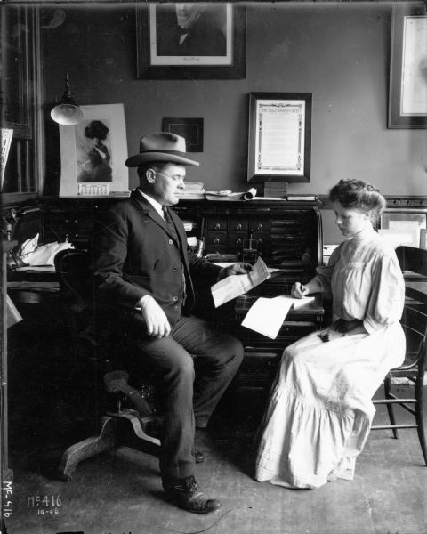 A businessman in a three-piece suit and hat sits behind a large desk with a paper in his hand, while a secretary in a long, high-collared dress writes on a pad. On the wall are a 1906 calendar and a framed piece of writing titled "The Salesman's Creed." The man may be the factory superintendant or some other manager at International Harvester's McCormick Works in Chicago.