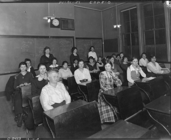Women pose at their desks in an evening class.  Three women (teachers?) stand in the back.  Mathematical equations are written on the blackboards in the back of the room. The women are likely employees or prospective employees at International Harvester's Osborne Works (later known as Auburn Works) in Auburn, New York.