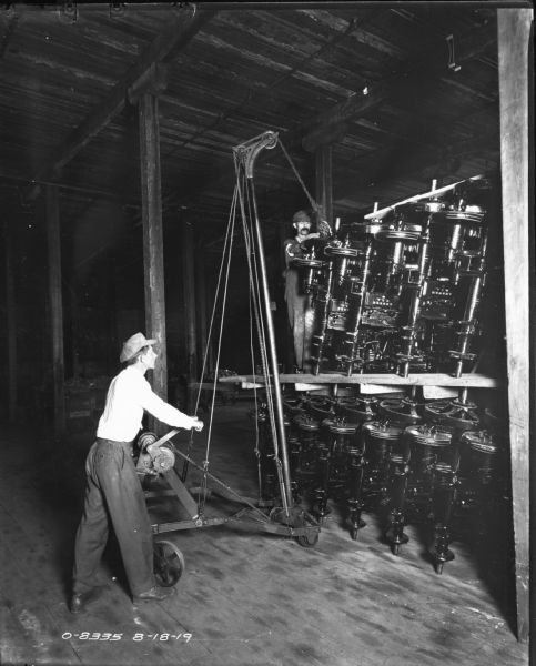 Two men use a crane to move large parts inside an International Harvester manufacturing plant. The factory is likely International Harvester's Osborne Works (later known as Auburn Works) in Auburn, New York. The parts are stamped Auburn U.S.A.