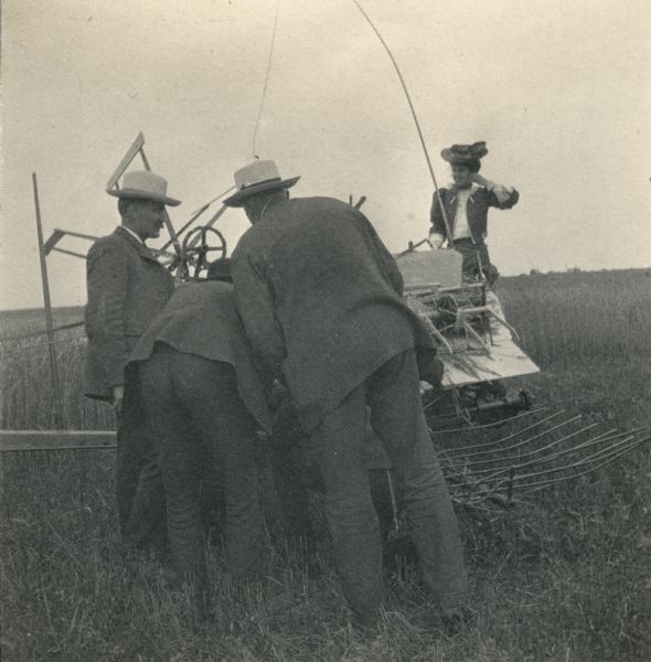 Three men in suits work on an International Harvester grain binder while Katherine Dexter McCormick sits in the seat of the machine. The man bent over the machine in the center is likely a Mr. Cavanaugh. The other two men are two of the following: Mr. Pridmore, Mr. Brooks and/or Mr. Sharp of the Osborne Experimental Department. The photograph was likely taken by Katherine McCormick's husband Stanley McCormick.