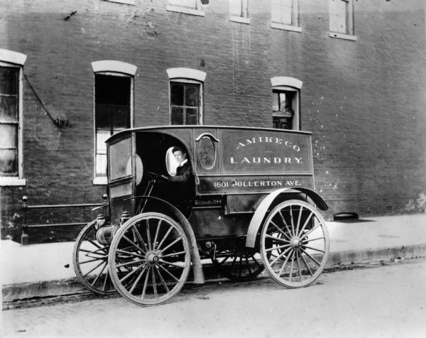 A man drives a 1909 International Auto Wagon belonging to the Amikeco Laundry at 1601 Fullerton Avenue in Chicago. The truck has an enclosed body and crude fenders.