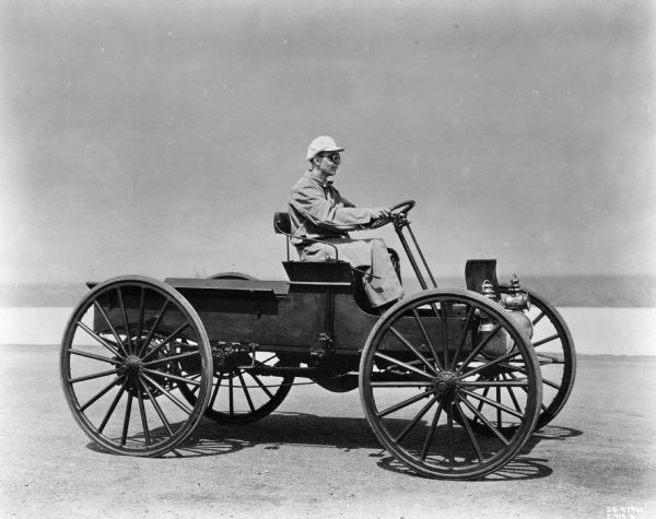 A man wearing goggles, a hat and a duster is driving an "antique" 1909 International Auto Wagon.