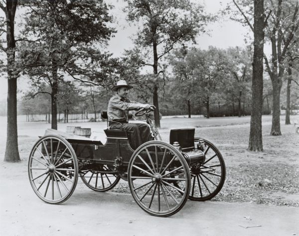 A man in a hat drives a 1909 International Auto Wagon loaded with lumber, barrels and a sack near a park or rural area.