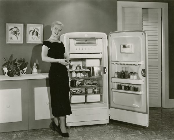A woman in a black dress and heels stands in a kitchen next to an International Harvester refrigerator stocked with food and beverages.