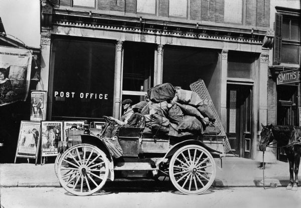 International Auto Wagon parked in front of a horse outside a post office. The truck is loaded with sacks, possibly filled with mail.