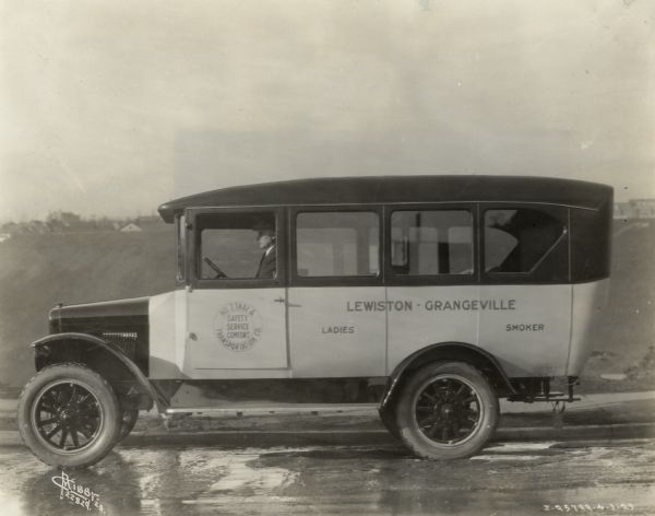 International truck serving as a bus or taxi, most likely carrying passengers between Lewiston and Grangeville, Idaho. The truck has "Lewiston-Grangeville Ladies Smoker" painted on the side. The driver, wearing a suit and cap, is behind the wheel.