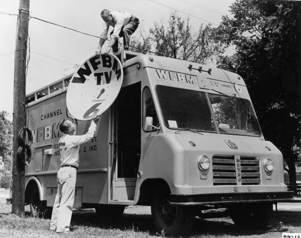 Two workers for WFBM-TV in Indianapolis attaching a satellite dish to their International AM-152 truck. The truck was used for remote television pickups. It accommodated a full four-man control crew, five cameras, cameria tripods, microphones and other TV equipment.